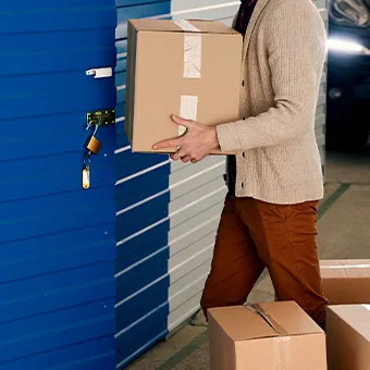 A man in a beige cardigan carrying a cardboard box past a blue storage unit with a padlock on its latch
