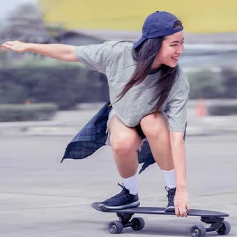 A smiling young woman in a cap and a grey T-shirt crouching on a moving skateboard with one arm stretched out