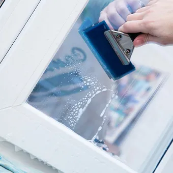 A hand pressing a blue squeegee across a wet window pane in a white frame