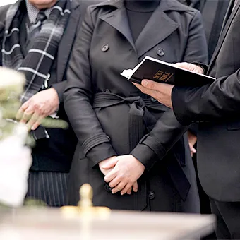 Mourners in dark coats standing beside a coffin, one holding an open Bible