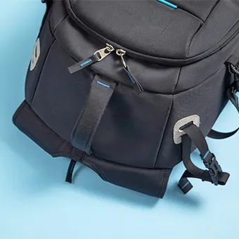 The lower half of a black backpack with blue trim, showing its zip pulls and side straps, on a pale blue background