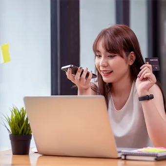 หญิงสาวยิ้มอยู่หน้าแล็ปท็อป มือหนึ่งถือโทรศัพท์ อีกมือถือบัตร มีกระถางต้นไม้เล็ก ๆ วางอยู่ข้าง ๆ