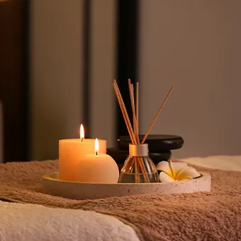 A tray with 2 lit candles, a reed diffuser, stacked black stones and a frangipani flower, resting on brown towels