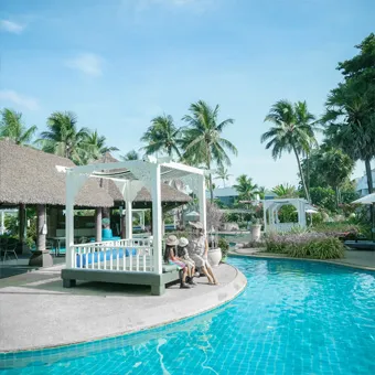 A woman and a child sitting in a white gazebo on a pool deck, with palm trees and thatched roofs behind