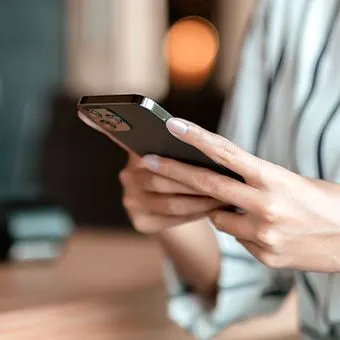 A close-up of a hand with pale nails holding a phone in front of a blurred room