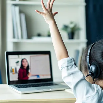 A child in headphones raising a hand towards a laptop screen that shows a woman in a red top in front of a whiteboard