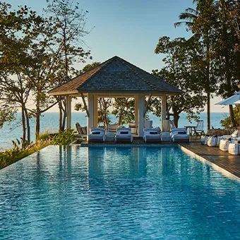 A swimming pool with a pavilion and white sun loungers at its far end, with trees and the sea behind it in late afternoon light