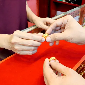 มือหนึ่งยื่นแหวนทองให้อีกมือหนึ่งข้ามตู้กระจกที่ปูผ้าสีแดง ด้านล่างมีอีกมือถือแหวนทองอีกวง