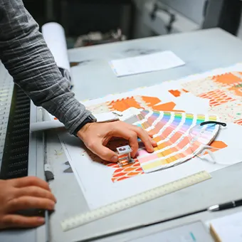 A hand spreading a fan of colour swatches across a printed proof on a work table, with a ruler beside it