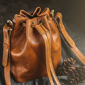 A tan leather bucket bag with a drawstring top beside pine cones on a dark cloth