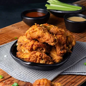A black bowl of fried chicken on a wooden board, with dipping sauces and celery sticks behind