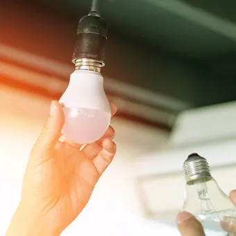A hand screwing a white bulb into a ceiling holder while another hand holds a clear glass bulb
