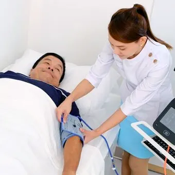A woman in a white tunic wraps a blood pressure cuff around the arm of a man lying in bed under a white blanket