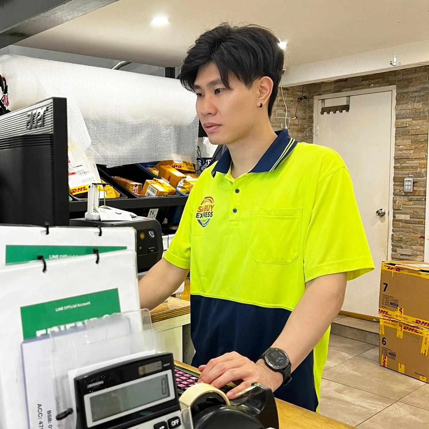 A man in a yellow-green and navy polo shirt with a Sabuy Express logo stands at a desk working at a computer, with parcels on shelves behind him