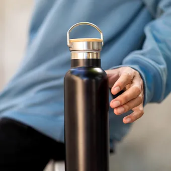 A black steel bottle with a bamboo lid and a metal carry handle, held by a person in a light blue sweatshirt