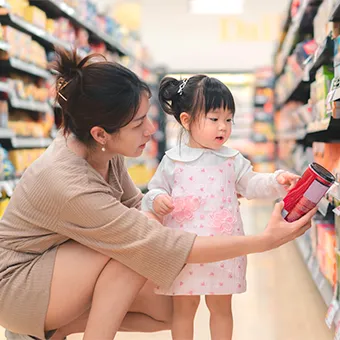 A woman crouching beside a small girl in a pink dress as they look at a red tin in a supermarket aisle