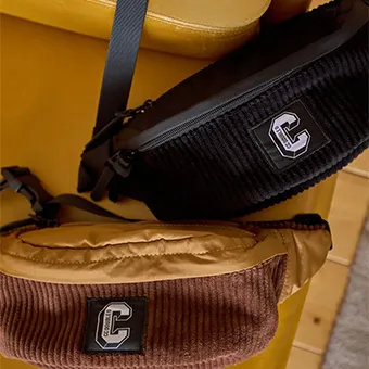 Two corduroy waist bags with a white letter C patch, one black and one brown with a mustard top, hanging on a yellow chair