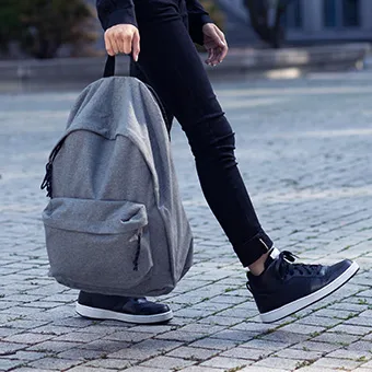 A person in black jeans and dark trainers walking on paving stones, carrying a grey backpack by its top handle
