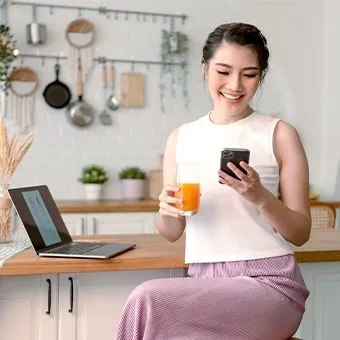 A smiling woman holding a phone and a glass of orange juice at a kitchen counter, beside an open laptop