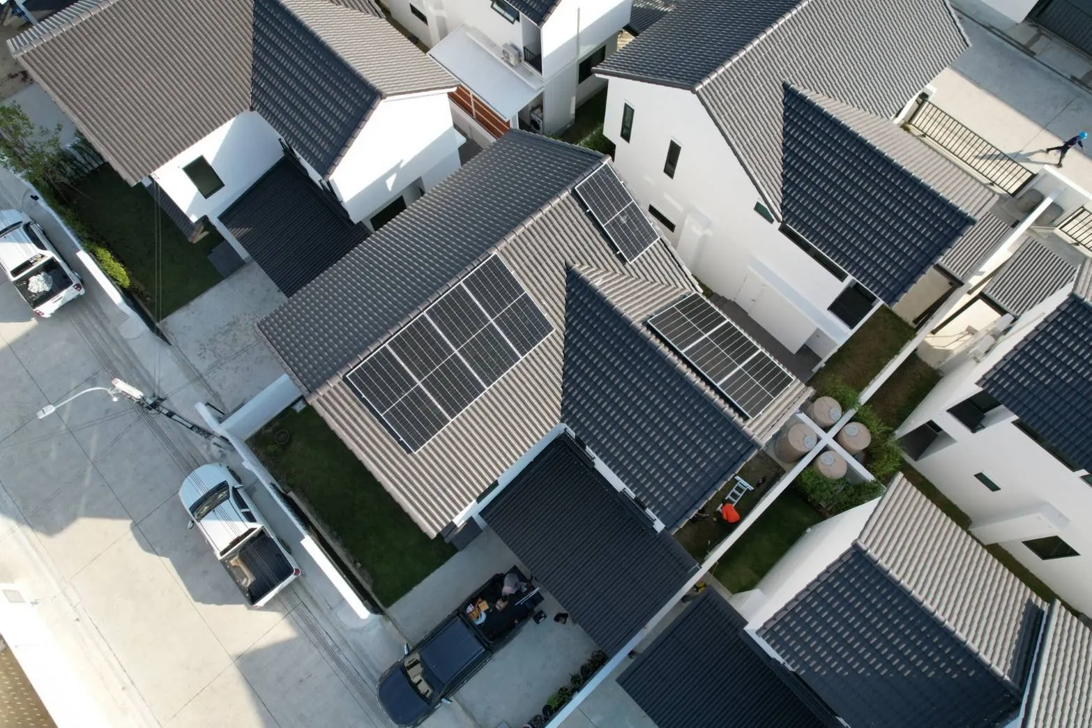 Aerial view of a housing estate, with 3 groups of solar panels on the grey tiled roof of one house among white houses with dark roofs and pickup trucks parked nearby