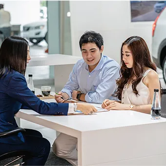 A woman in a blue suit going through papers with a smiling man and woman at a white desk in a car showroom