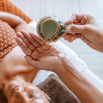 Hands pouring oil from a small patterned pot into an open palm above a person lying on a treatment bed