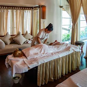 A therapist giving a massage to a guest lying under a white sheet on a massage bed, in a room with curtained windows