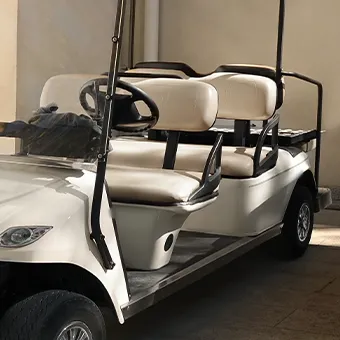 A white golf cart with 2 rows of cream seats parked in the shade beside a wall
