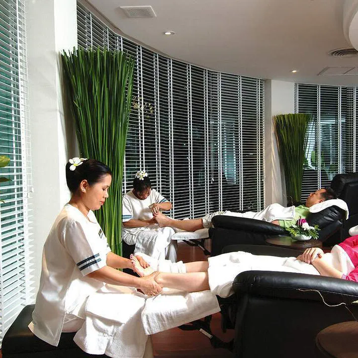 Therapists in white uniforms giving foot massages to 2 guests in reclining chairs beside tall green plants