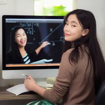 A girl at a desk with a pen in her hand looking back over her shoulder, a computer screen behind her showing a woman at a board of maths
