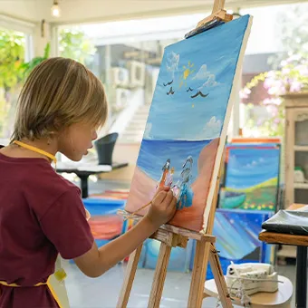 A child painting a beach scene on a canvas on an easel in a bright art room