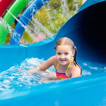 A smiling girl in a rainbow swimsuit sliding into the water at the end of a blue slide