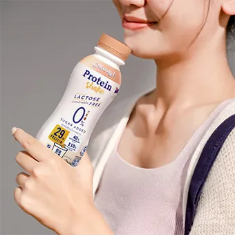 ผู้หญิงเสื้อสีม่วงอ่อนยิ้มพร้อมถือขวด Hooray Protein Shake ไว้ใกล้ใบหน้า