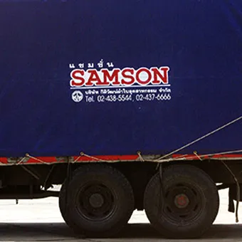 ผ้าใบสีกรมท่าบนรถบรรทุกพิมพ์คำว่า SAMSON สีแดงขอบขาว มีคำว่า แซมซั่น อยู่ด้านบน และชื่อบริษัทกับเบอร์โทรศัพท์ 2 หมายเลขอยู่ด้านล่าง เหนือล้อหลังของรถ