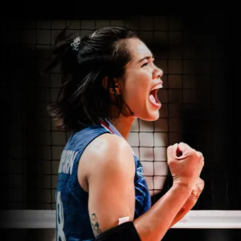 A woman volleyball player in a blue shirt cheering with her mouth open and her fist clenched in front of the net