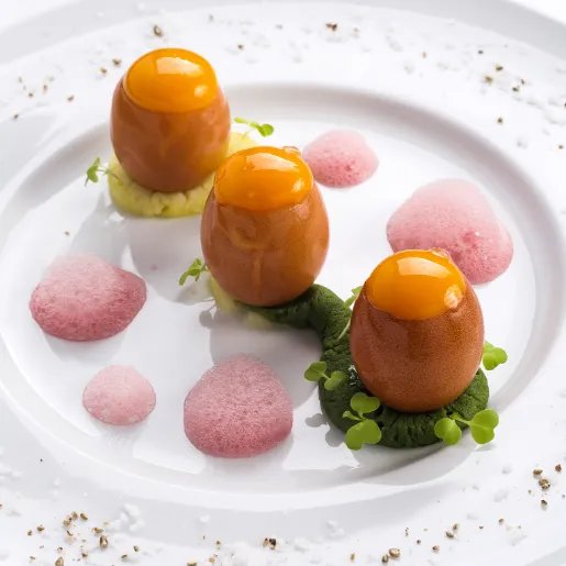 Three round bites with orange tops on green and yellow purée, with small mounds of pink foam, on a white plate