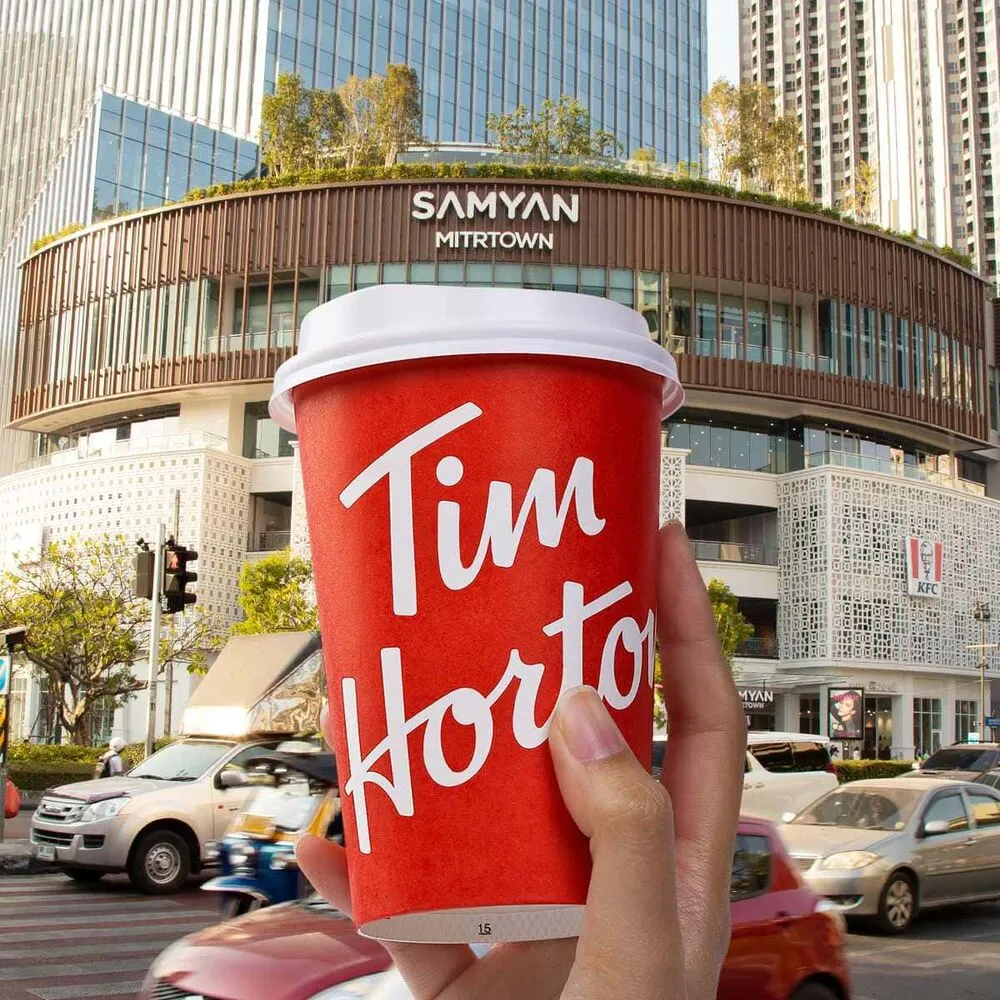 มือถือแก้วกาแฟสีแดงของ Tim Hortons หน้าศูนย์การค้าริมถนนที่มีรถสัญจรในกรุงเทพฯ
