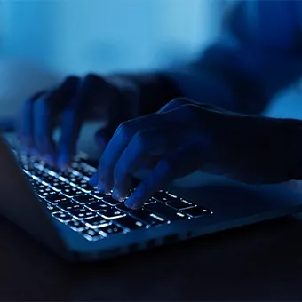 Hands typing on a backlit laptop keyboard in a dark room lit blue