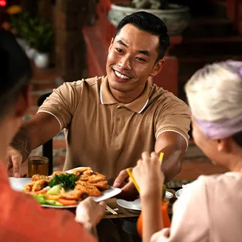 A smiling man in a polo shirt passing a plate of fried food and salad across a table to friends
