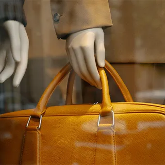 A mannequin’s hand holding the handles of a mustard yellow leather bag behind glass