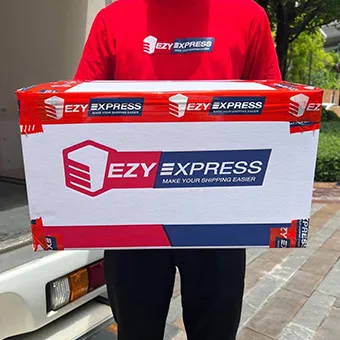 คนสวมเสื้อยืดสีแดงของ Ezy Express ถือกล่องพัสดุใบใหญ่ของ Ezy Express ที่ปิดด้วยเทปสีแดง