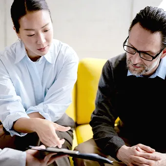 A woman pointing at a tablet as a man in glasses listens beside her on a yellow sofa