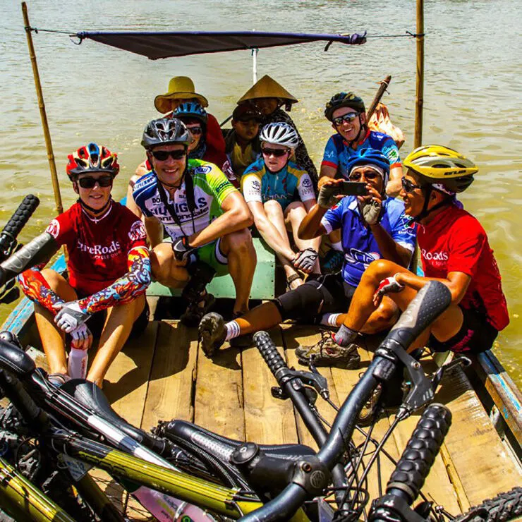 Cyclists in helmets and SpiceRoads jerseys sitting on a small wooden boat on a river, their bikes in front