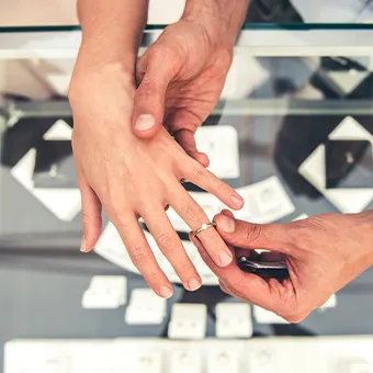 A person sliding a ring onto another person’s finger above a glass jewellery display case
