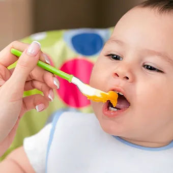 A hand feeding a baby orange purée from a green spoon