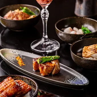 Glazed grilled meat with a green pepper on a long dark plate, with small bowls of food and a wine glass around it