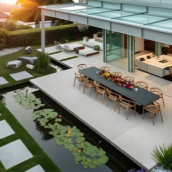 A long dining table with wooden chairs on a stone terrace beside a lily pond, next to a modern house with a glass roof