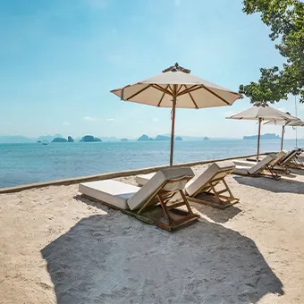 Wooden sun loungers under white parasols on a sandy beach, facing a calm sea with limestone islands on the horizon
