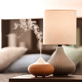 A wooden aroma diffuser letting out mist beside a table lamp with a ribbed grey base, on a bedside table