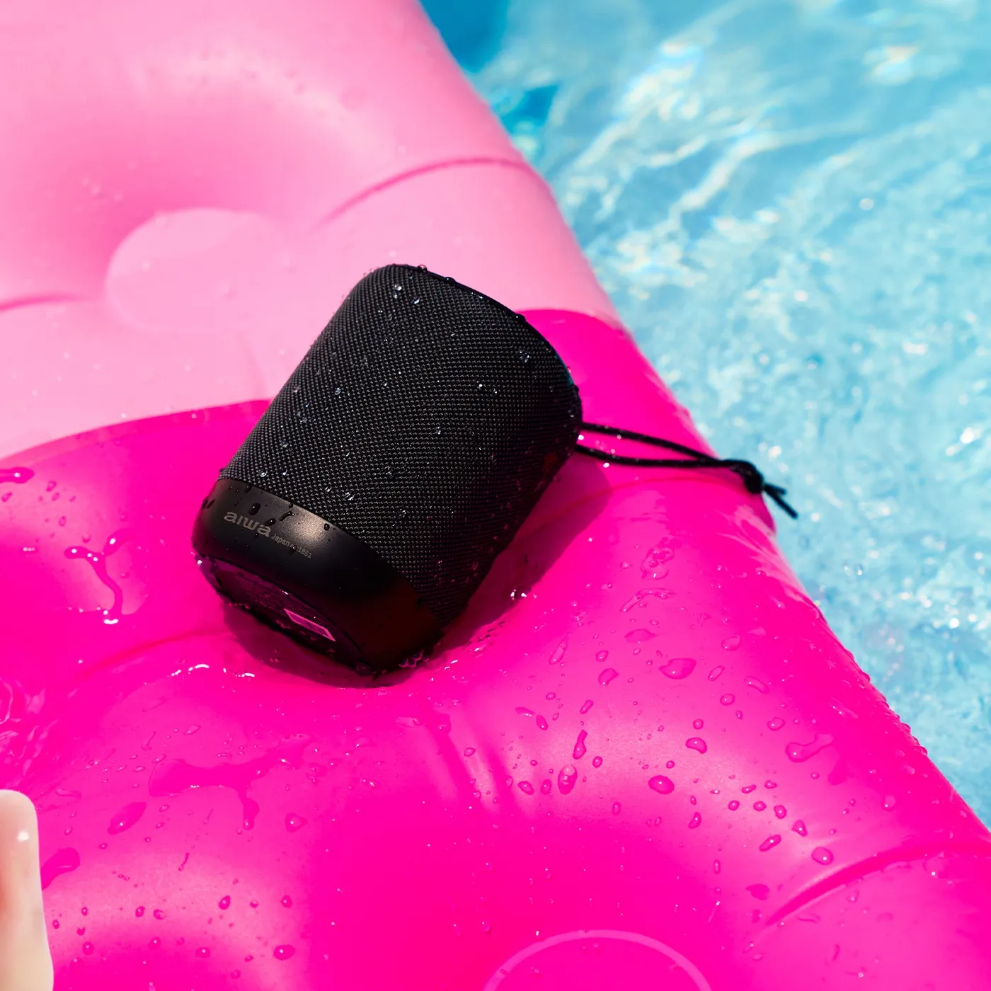A wet black AIWA speaker lying on a bright pink pool float next to blue water